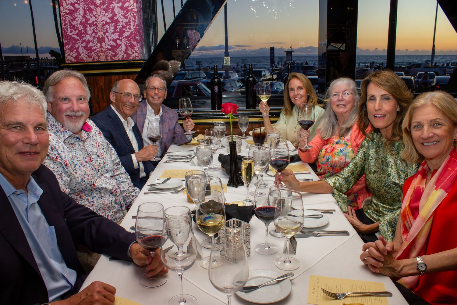 Group of eight adults enjoying a formal dinner party with wine glasses raised in a toast at a restaurant with city views.