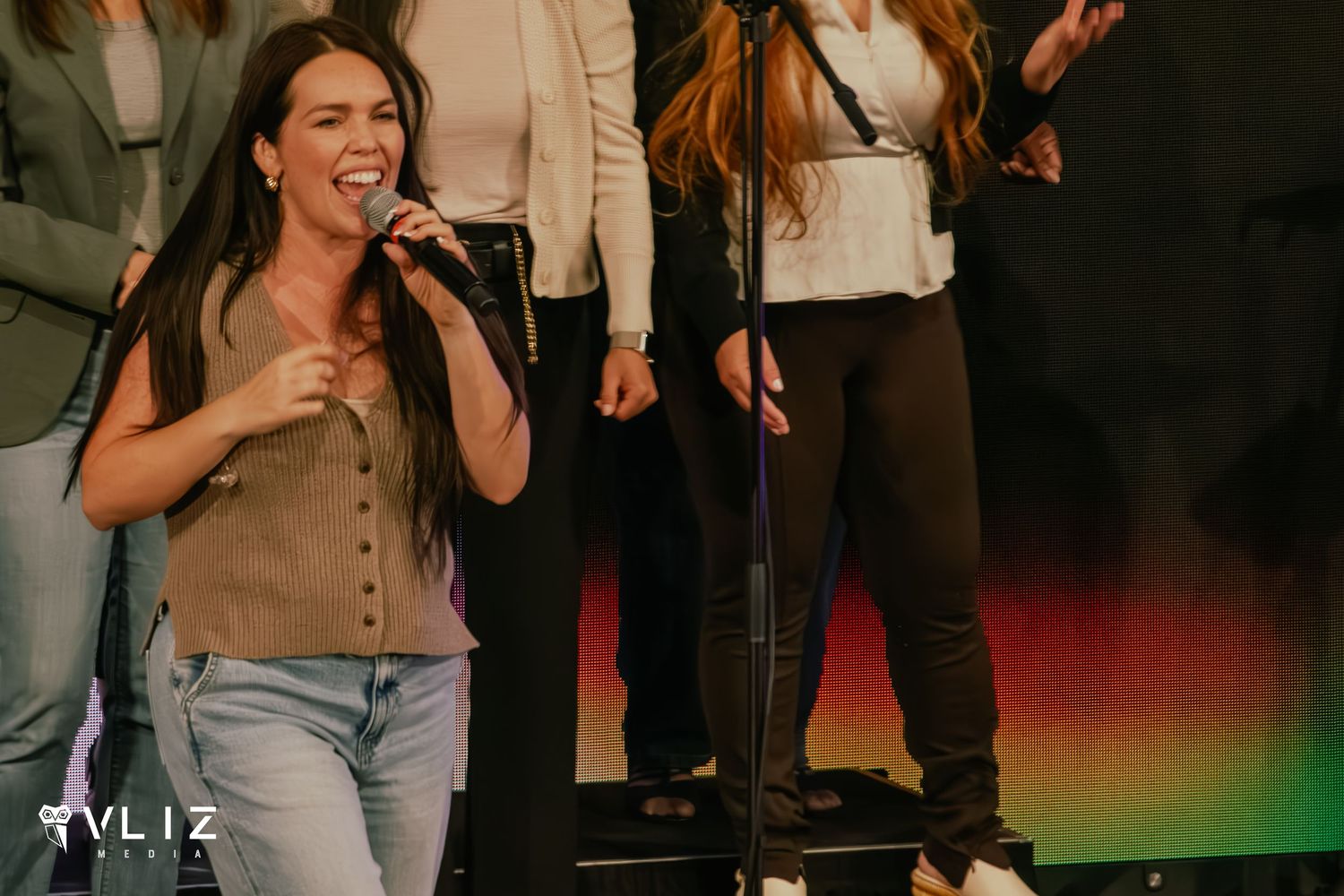 A woman passionately singing into a microphone on stage, wearing a tan top and jeans, with performers behind her.