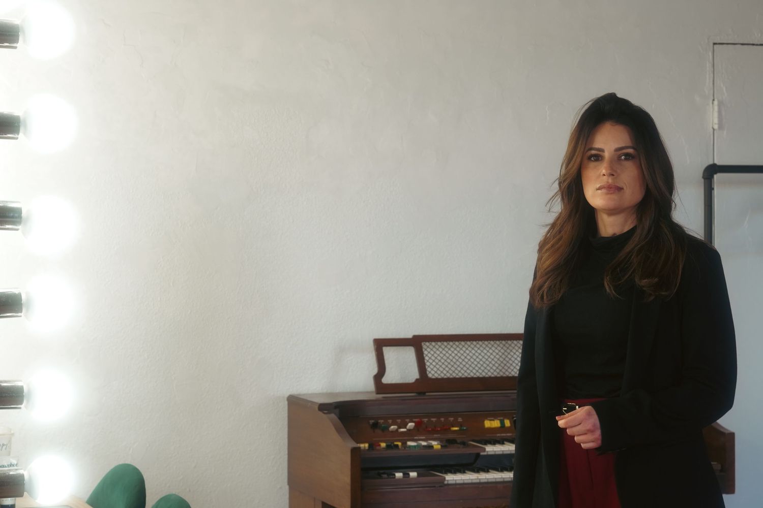 Woman in black coat standing near vintage organ with vanity mirror lights in a modern studio space.