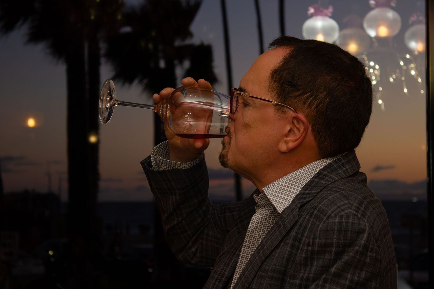 Man sipping red wine from a large glass at twilight with palm trees in background.