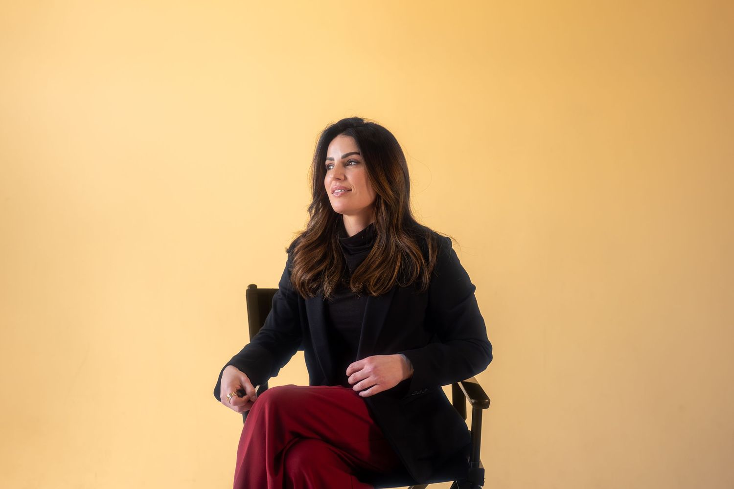 Woman in black blazer and red pants sitting on chair against yellow background, looking away thoughtfully.