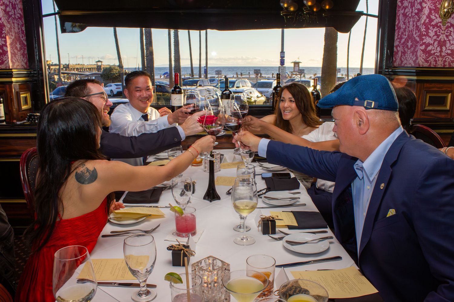 Group of six friends toasting with red wine glasses at a waterfront restaurant with harbor views.