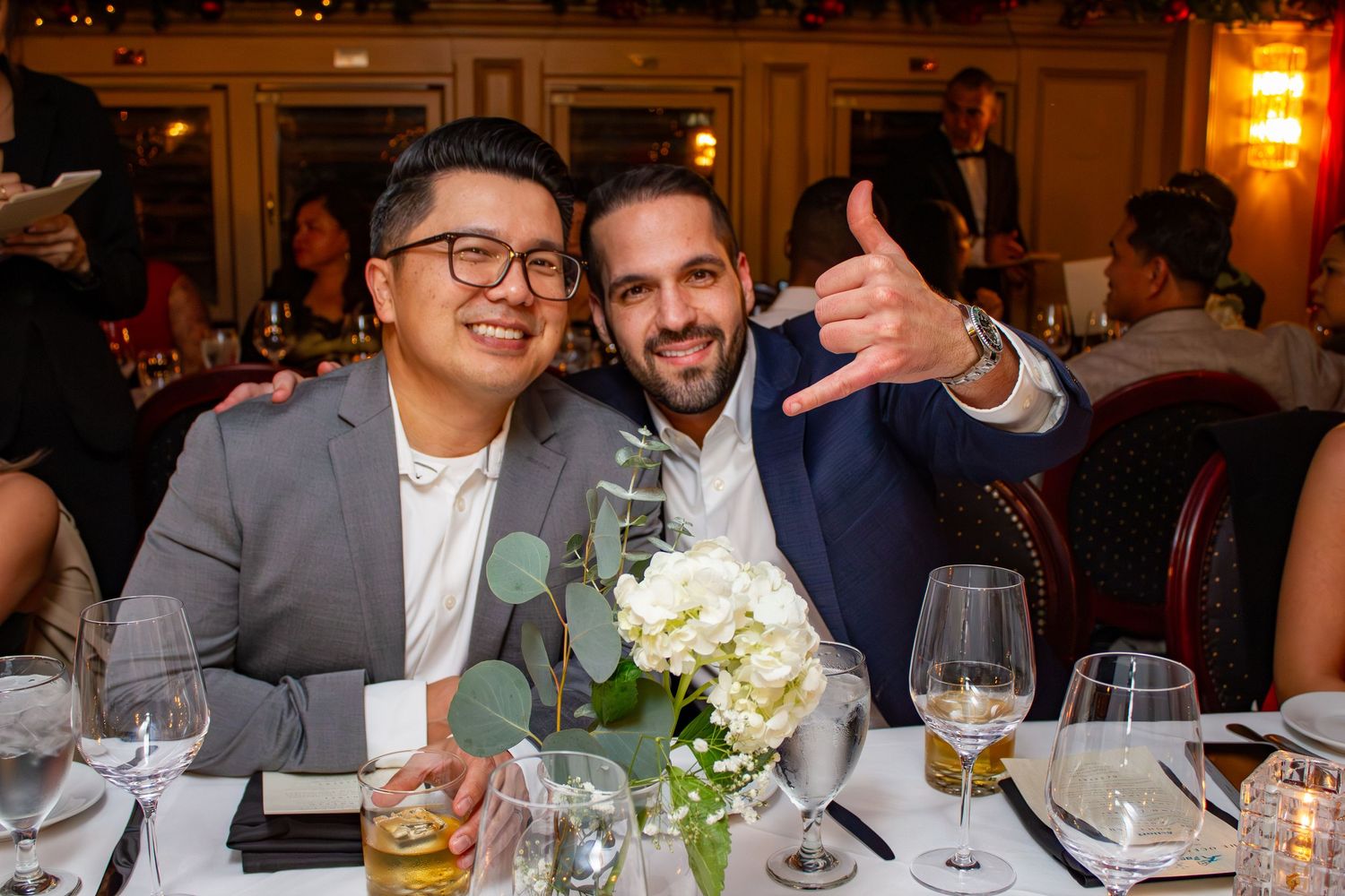 Two guests share a celebratory moment at an elegant dinner table with wine glasses and floral centerpiece.