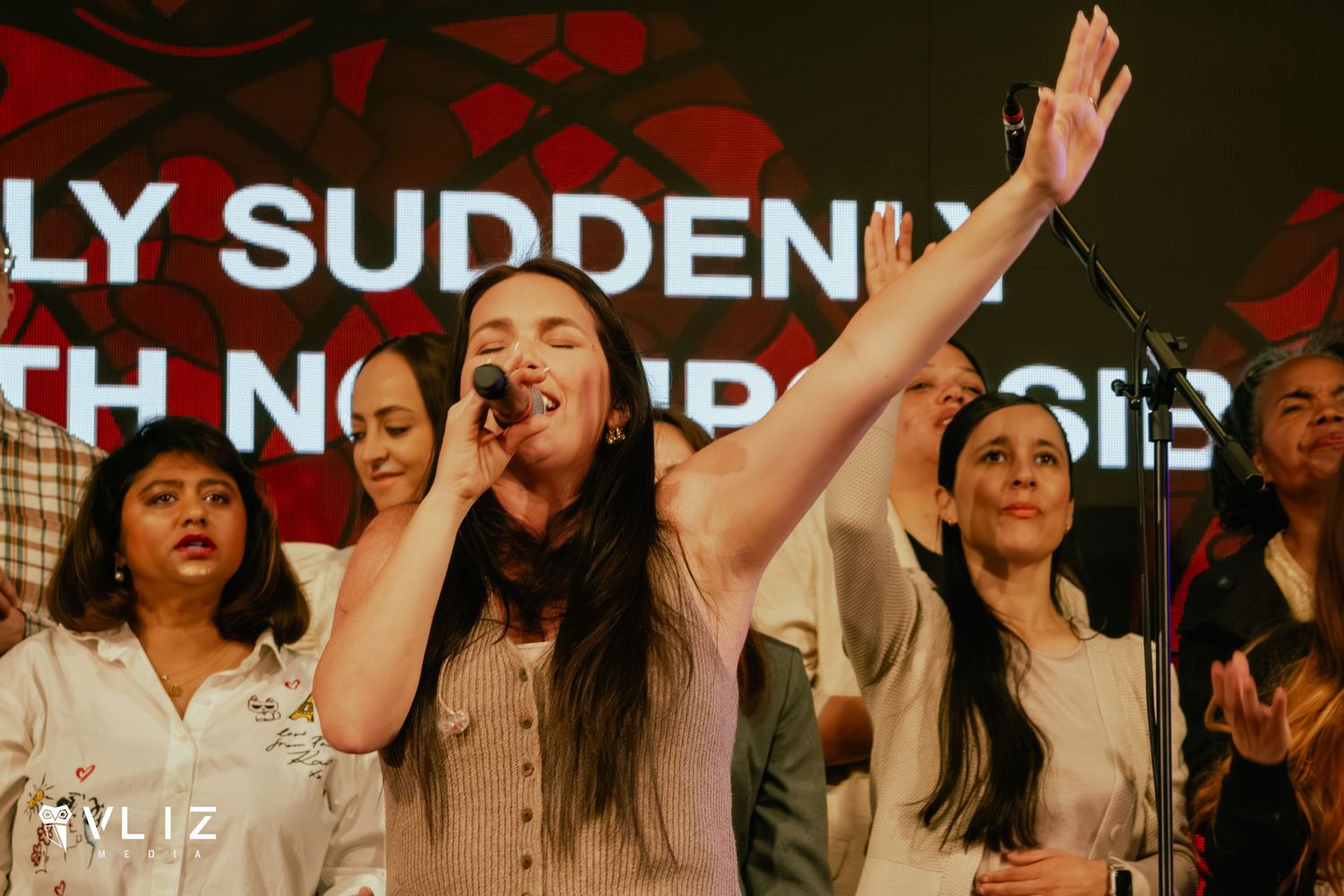 Woman singing on stage with raised arm leading choir at a live music event.