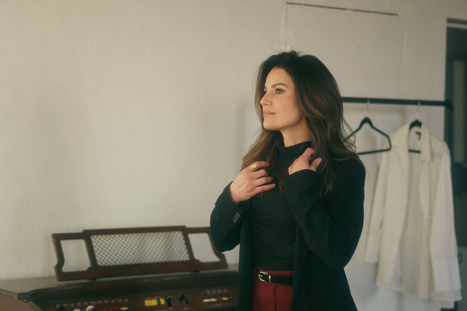 Woman in black top and red belt standing thoughtfully near a piano with white dresses on hangers in background.
