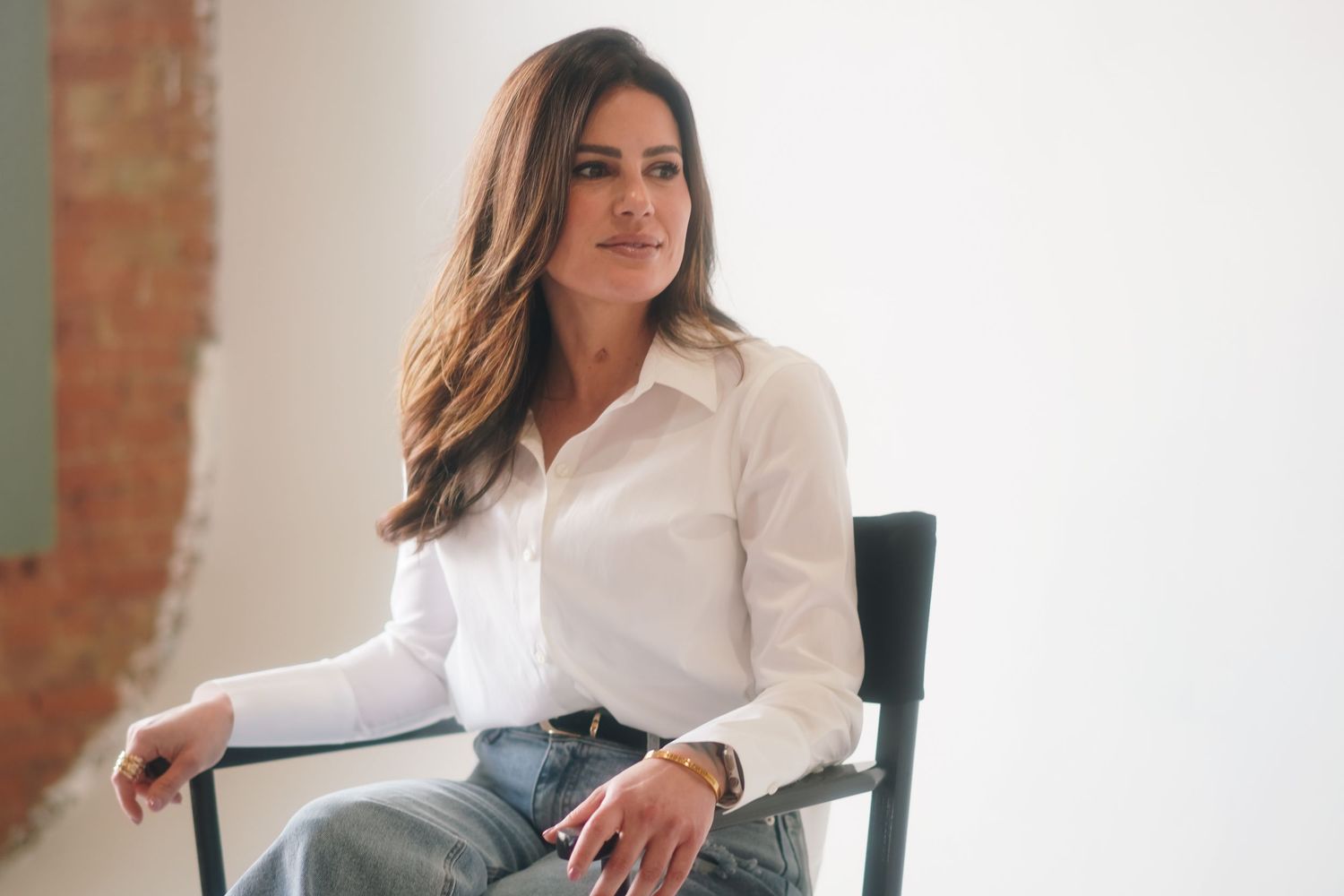 Woman in white button-up shirt and jeans sitting on dark chair against neutral background.