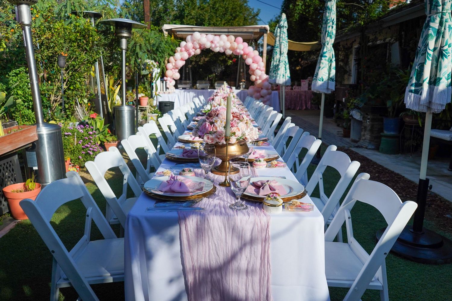 Long outdoor table set for a baby shower with pink decor and floral arch at the end.