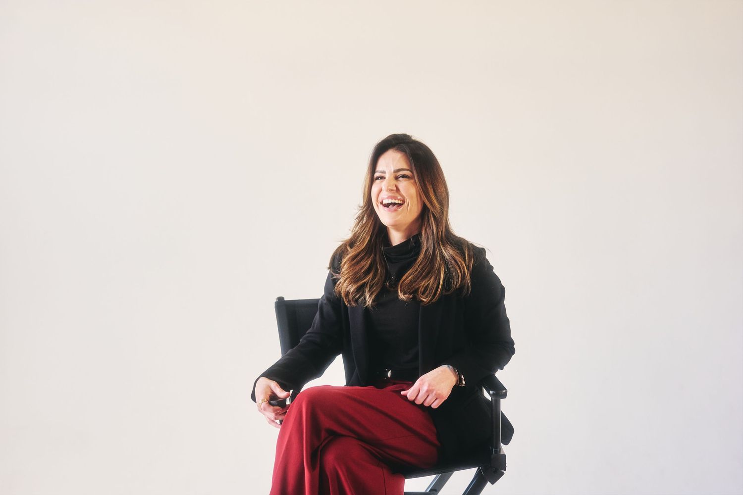 Woman in black turtleneck and red wide-leg pants laughing while seated on a director's chair against white background.