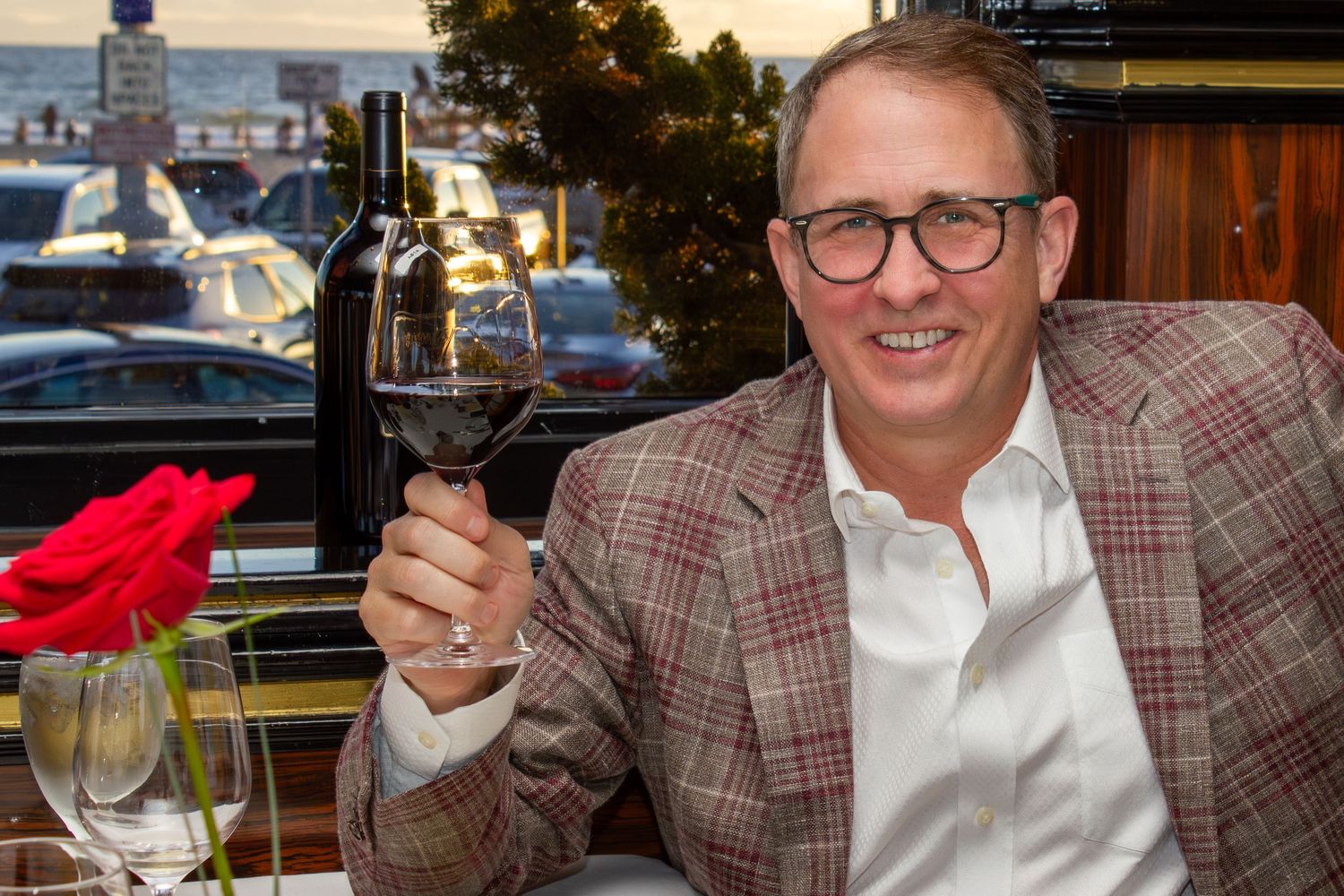 Man in plaid blazer toasting with a glass of red wine at a waterfront restaurant.