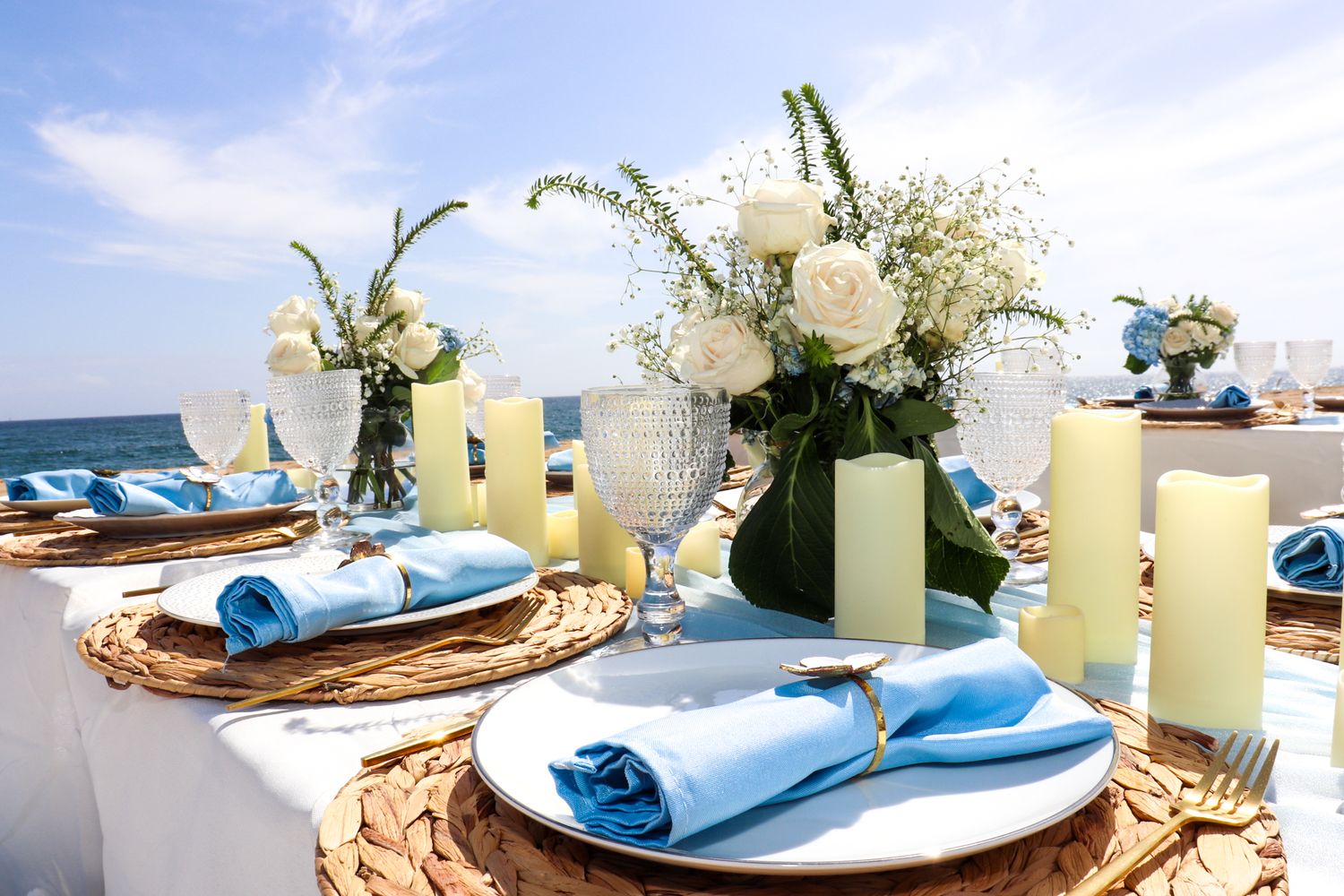 Elegant beach wedding table setting with blue napkins and white floral centerpieces.