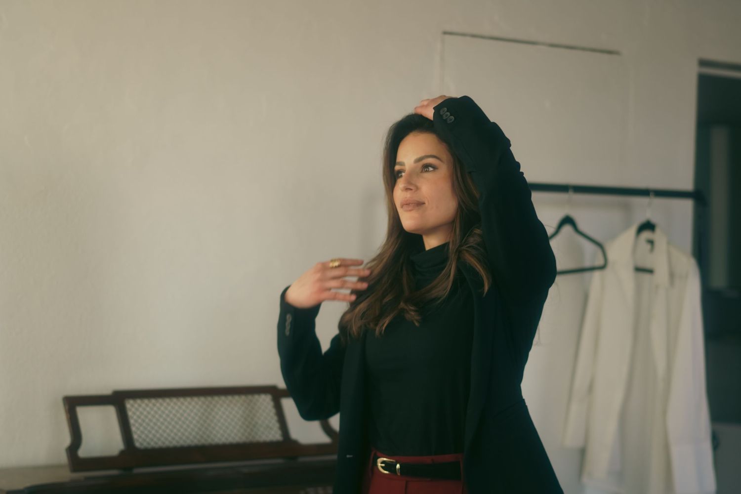 A young woman in a black outfit with a red belt poses thoughtfully in a bright room with white garments on hangers.