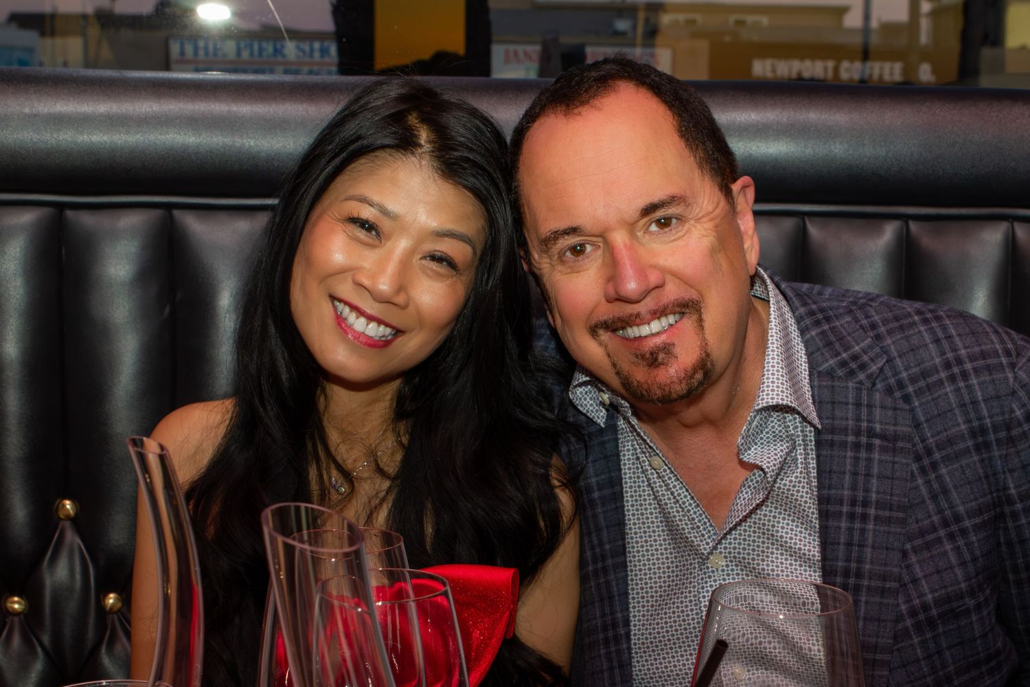 A smiling couple poses together at a restaurant, holding red drinks in a warmly lit setting.
