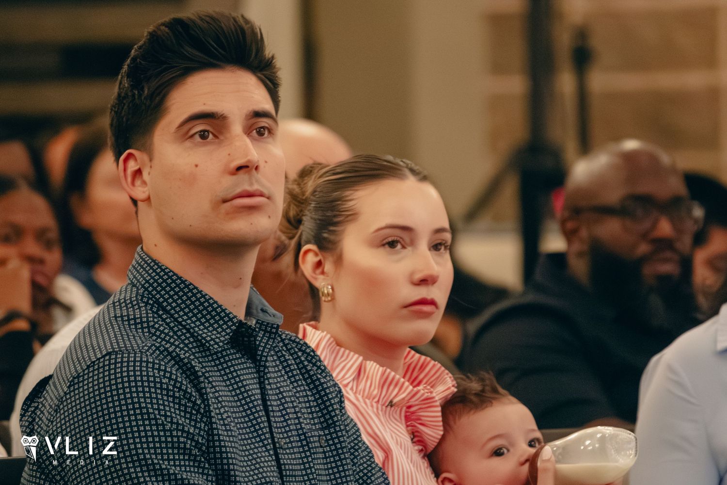 A man and woman sit attentively with a baby at an indoor event, looking serious.