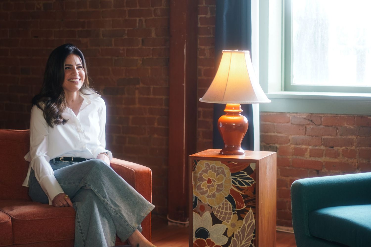 Woman in white blouse and jeans sits smiling in cozy brick-wall room with orange lamp.