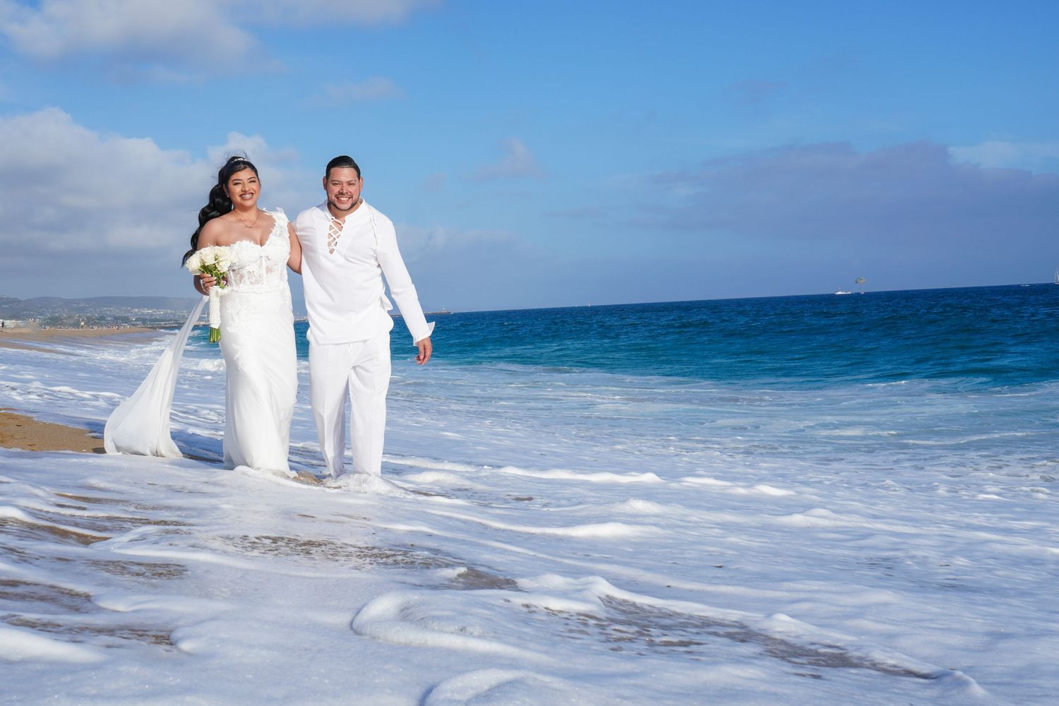 Couple walking along a scenic beach shoreline in white attire.