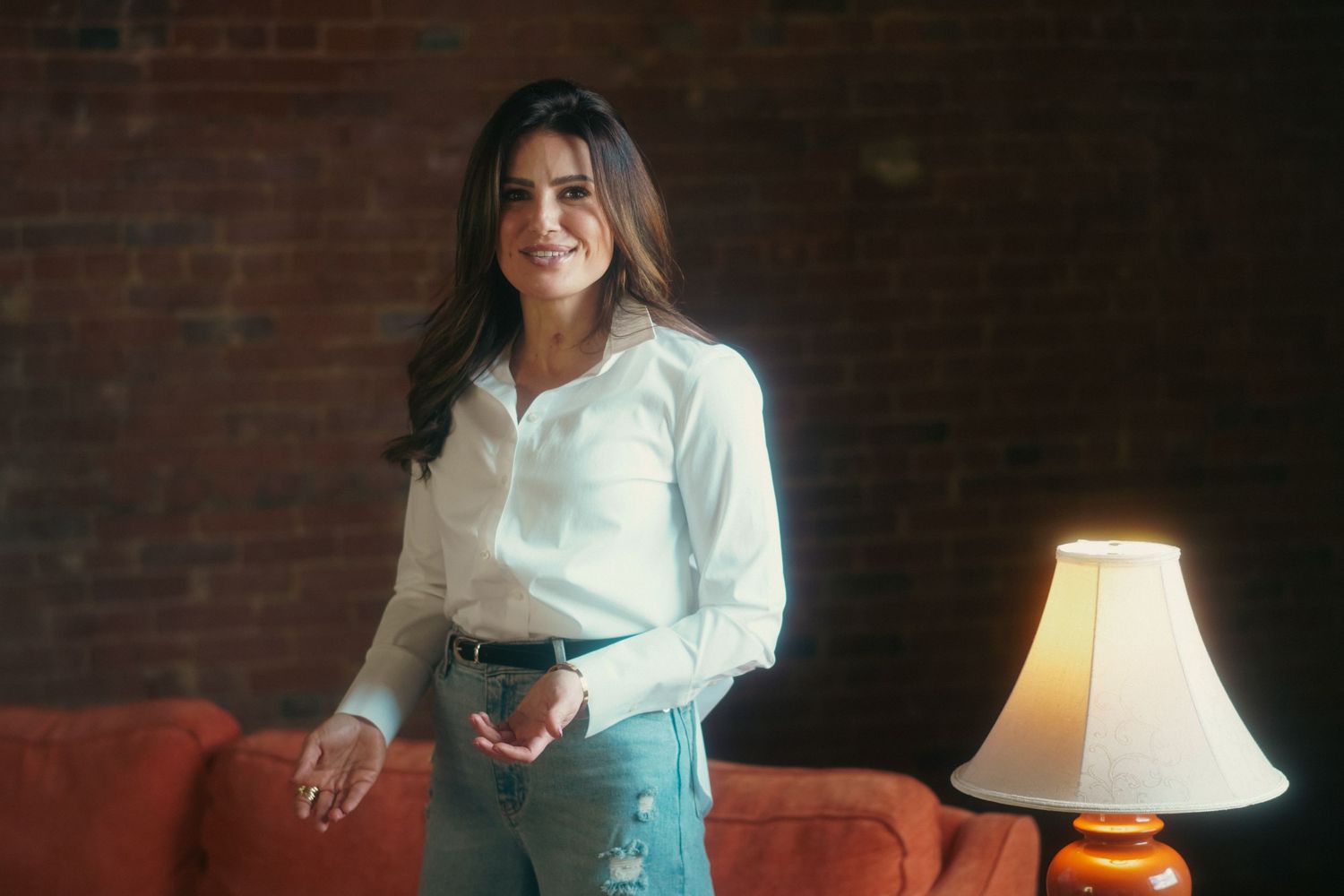 Woman in white shirt smiling confidently, standing near a red sofa and lamp against a brick wall.