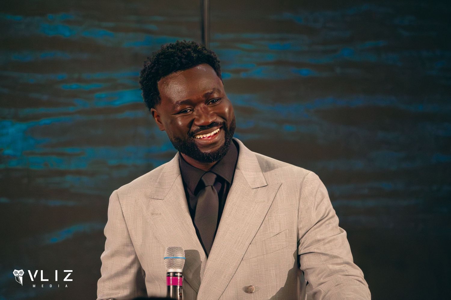 Smiling Black man in beige suit and dark tie seated at a panel discussion table with a microphone.