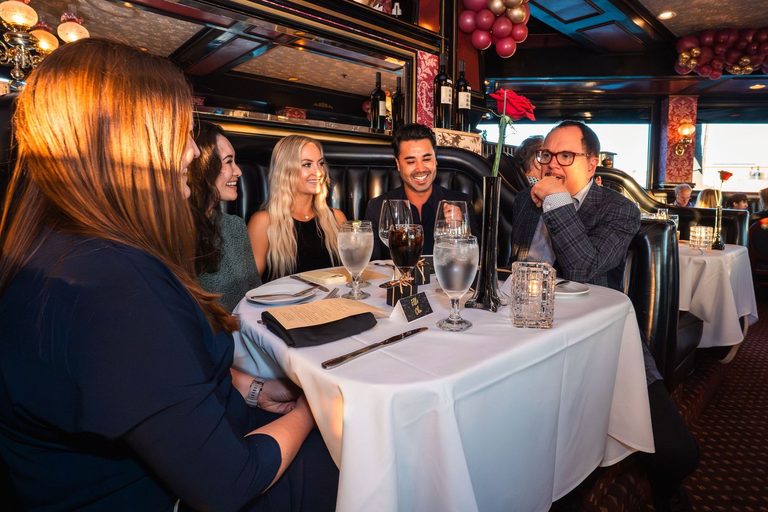 Group of friends laughing and enjoying dinner together at an upscale restaurant with white tablecloths.