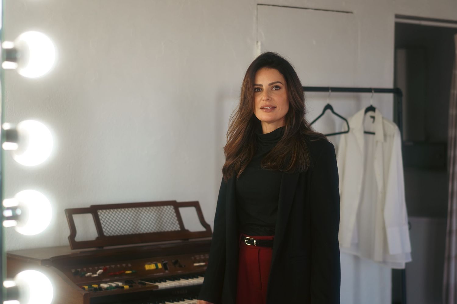 Woman in black coat and red belt stands in a dressing room with mirror lights and hanging white garments.