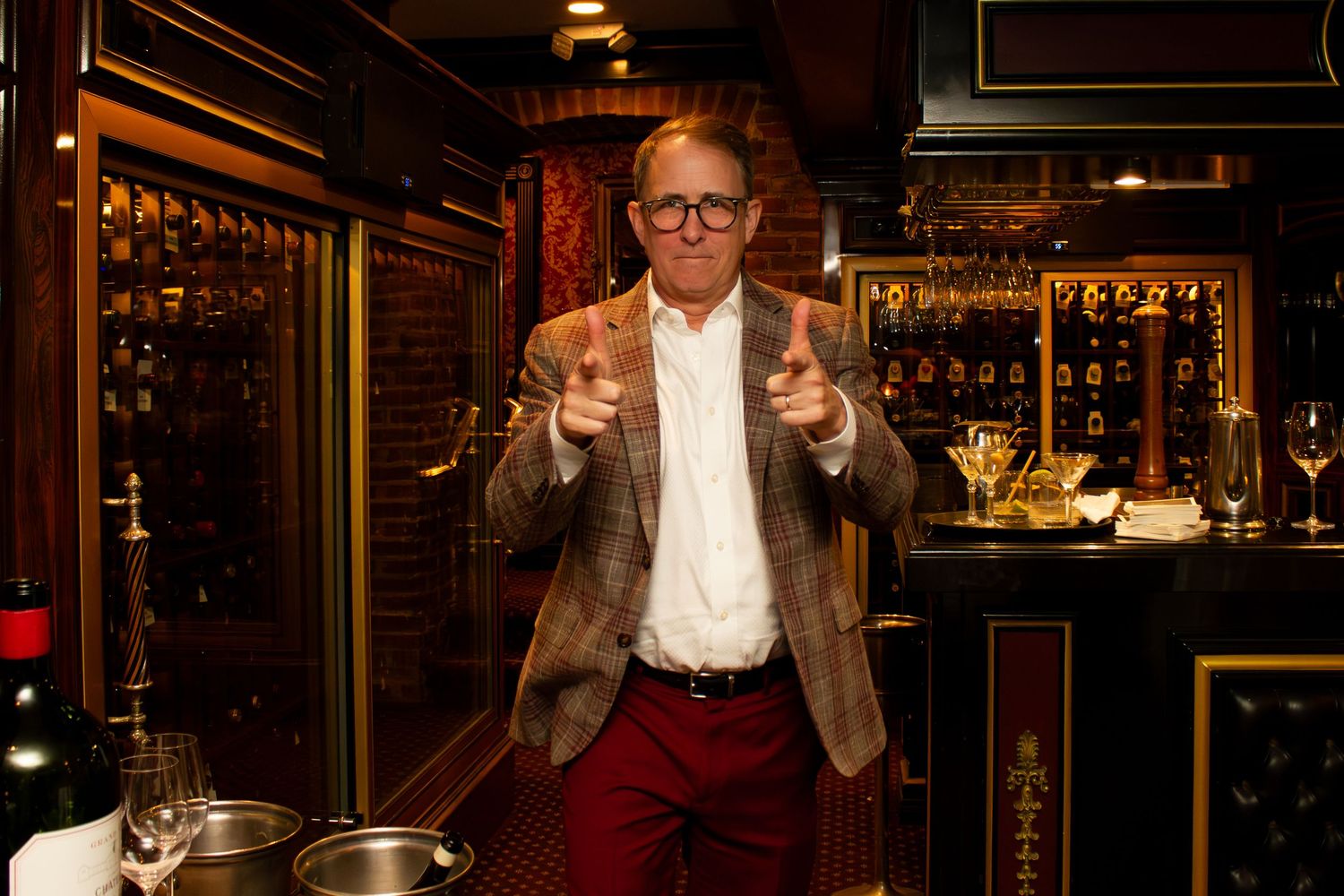Man in blazer and red pants pointing fingers in elegant wine bar with bottles and golden accents.