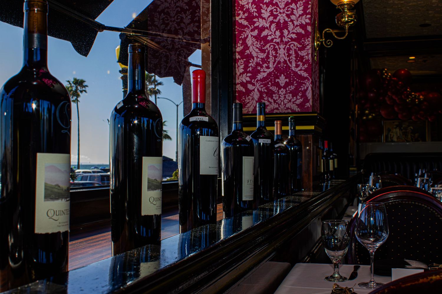 Row of dark wine bottles displayed on a reflective bar shelf in an elegant restaurant with red damask decor.