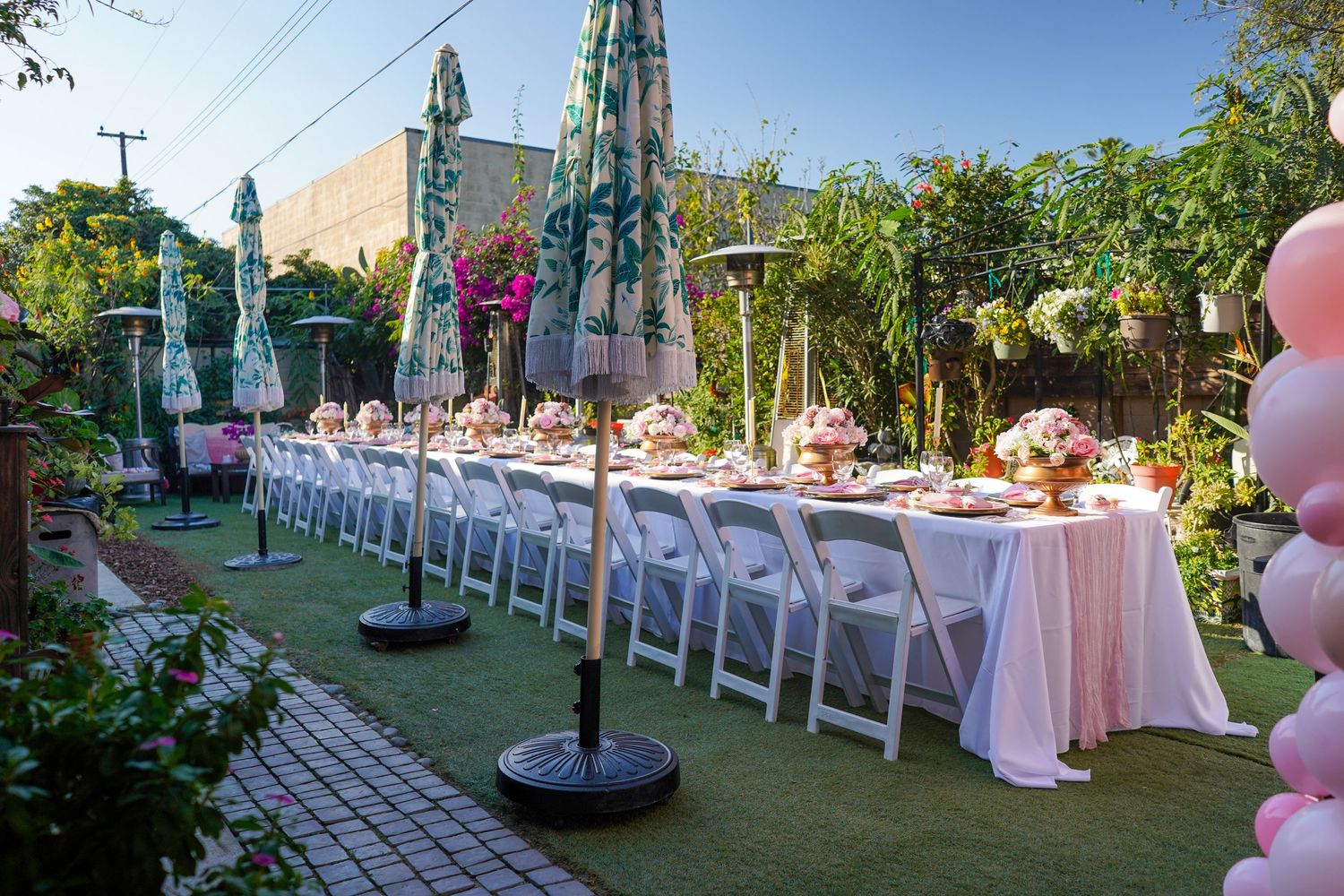 Long outdoor dining tables set up in a garden venue with white chairs and pink decor.