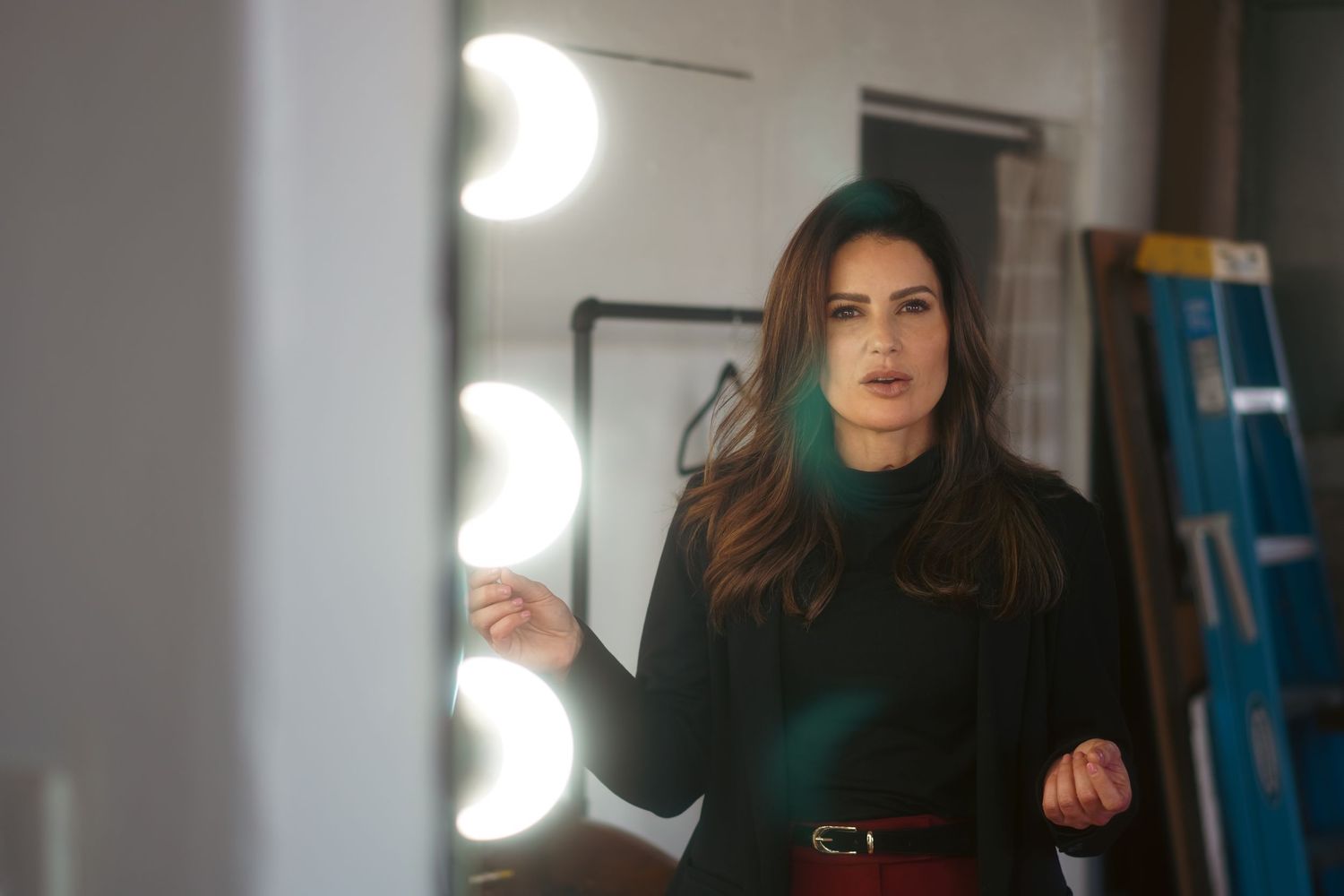 Woman in dark turtleneck and red belt gestures near vanity mirror with Hollywood lights in backstage setting.