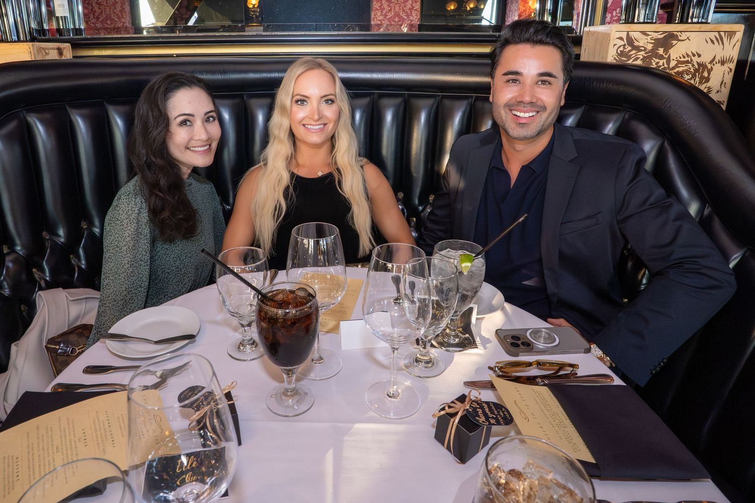 Three young adults smile while seated at a dining table in a upscale restaurant.