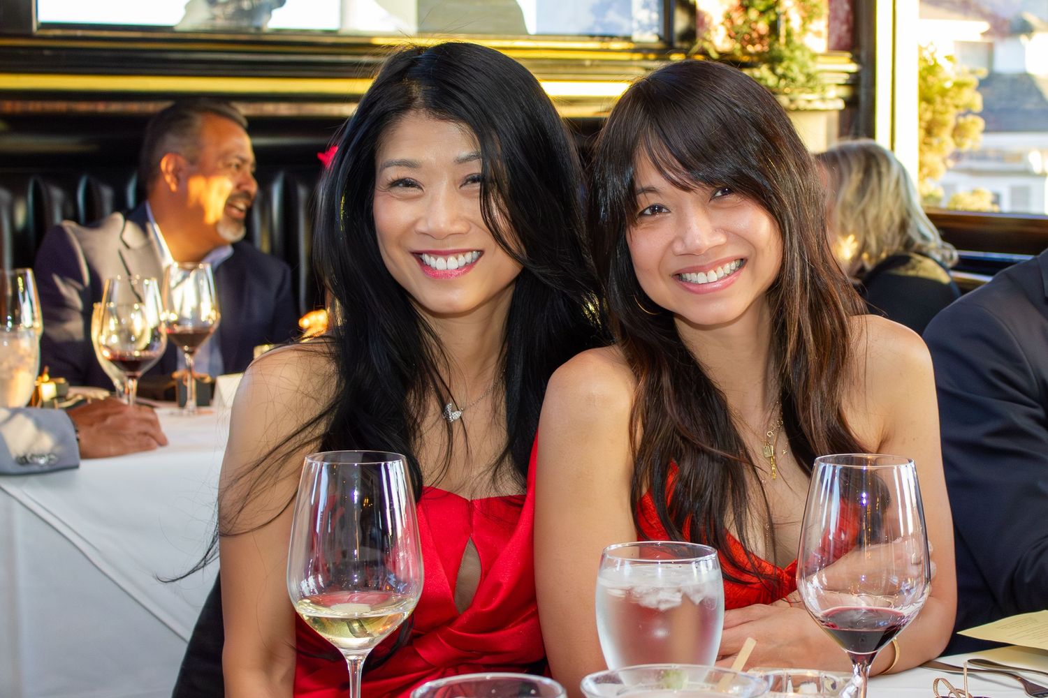 Two smiling Asian women in red dresses pose together at a restaurant with wine glasses.