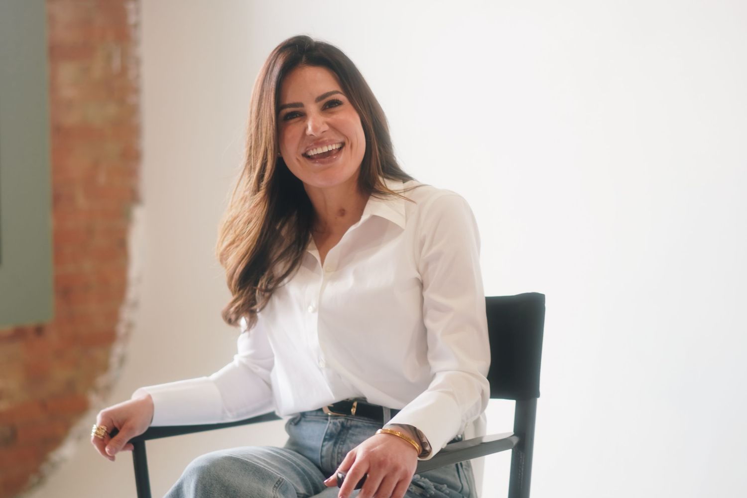Smiling woman in white button-up shirt and jeans sitting relaxed on a black chair against a white wall.