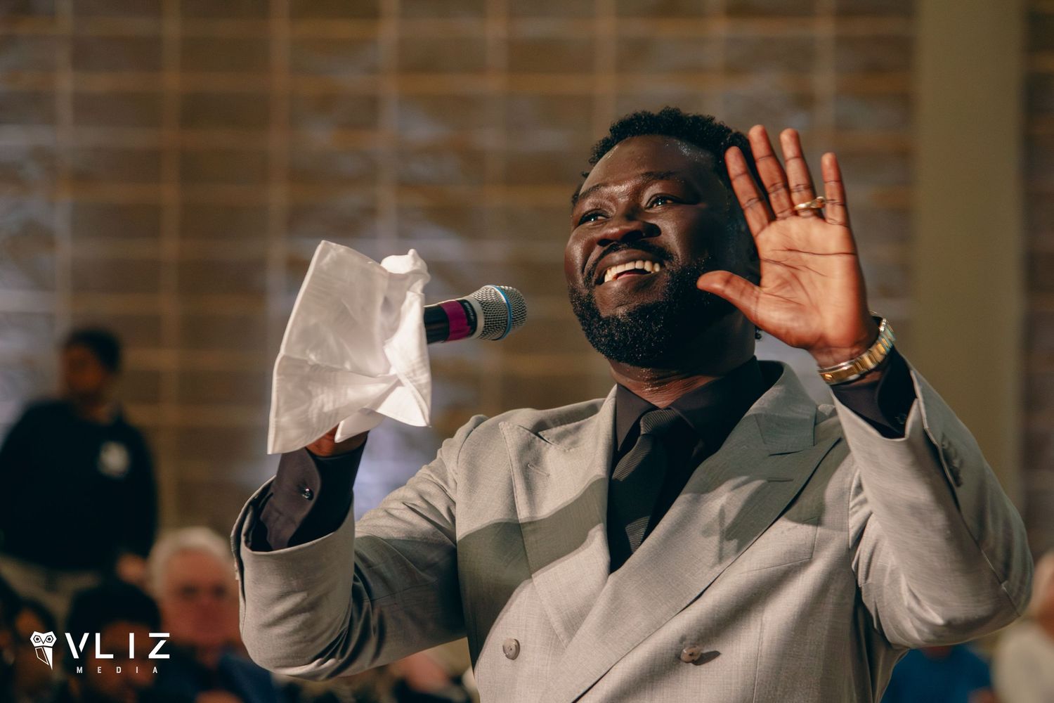 A joyful Black man in a gray suit sings into a microphone while raising his hand at an event.