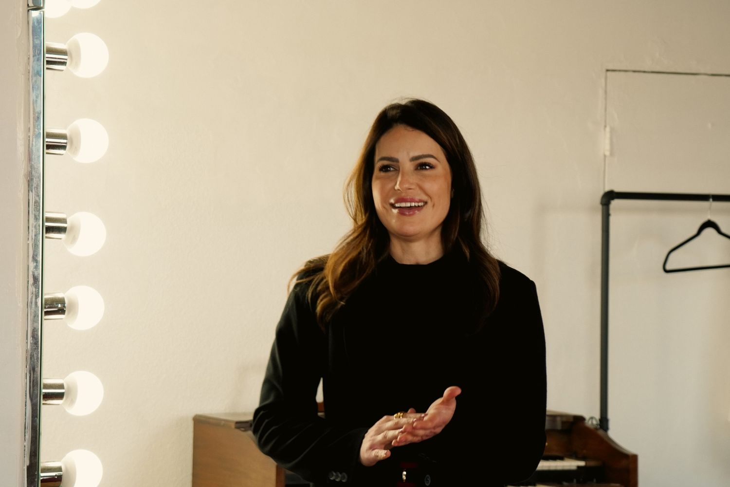 Woman in black top smiling in a dressing room with vanity lights and clothing rack in background.