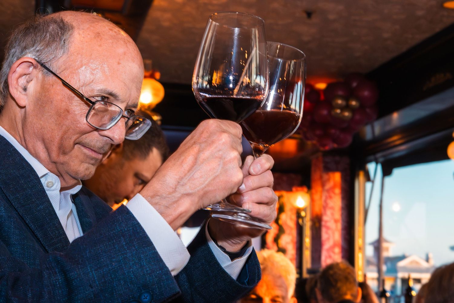An older man in a blue blazer carefully examines a glass of red wine at an upscale dining event.