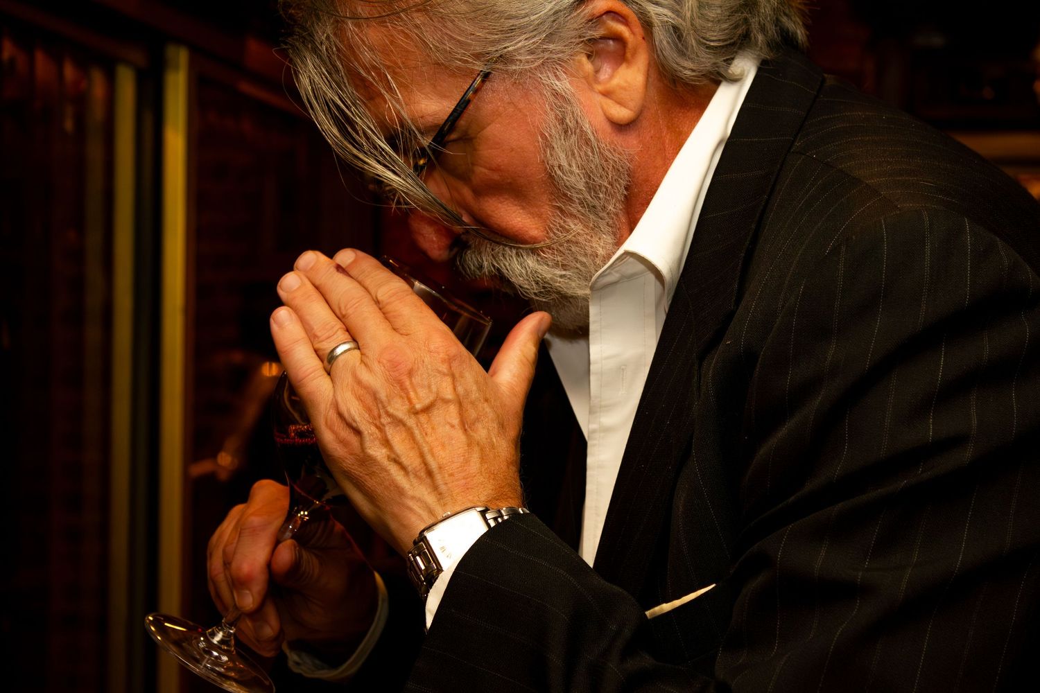 Elderly man with gray beard in dark suit sitting with hands clasped together in prayer or contemplation.