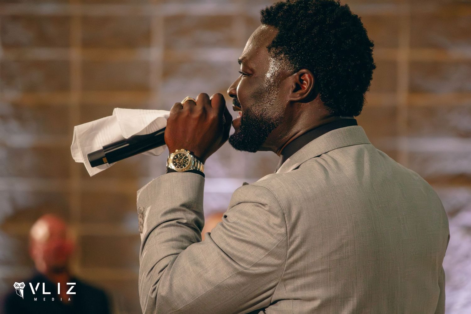 Man in beige suit singing into microphone at indoor event, wearing gold watch, performing on stage.