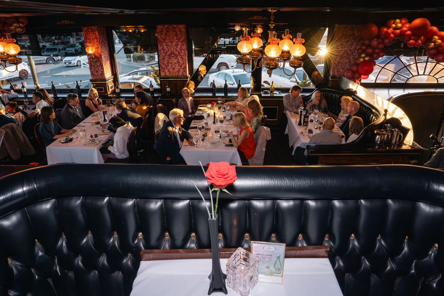 Elegant restaurant interior with black leather booths, white tablecloths, and a red rose centerpiece, filled with dining guests.