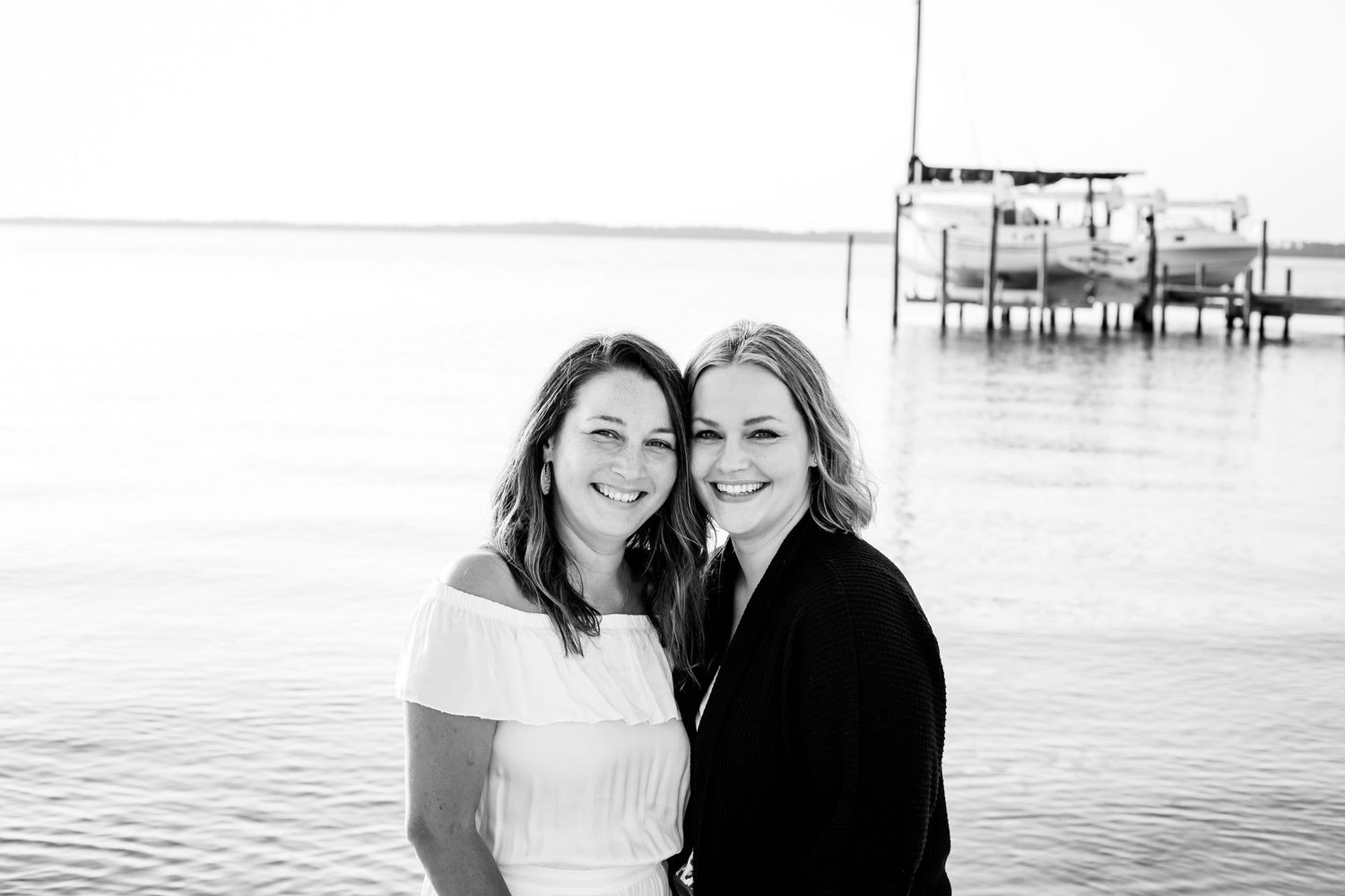 Black and white photo of two smiling women posing together near a waterfront dock outdoors.