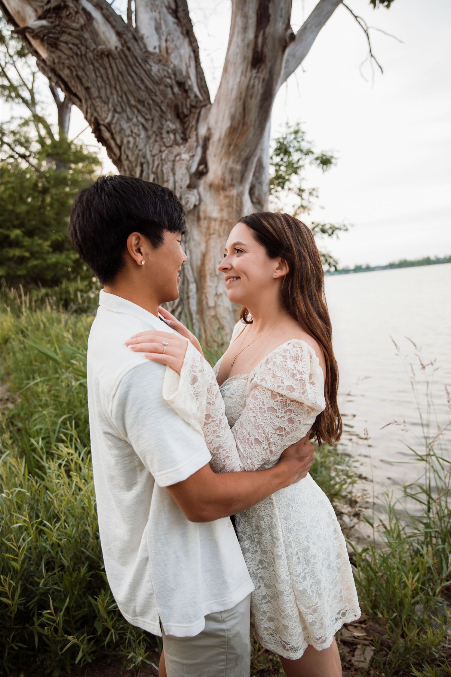 Smiling couple embracing near a large tree by a lake, gazing at each other in a romantic outdoor scene.