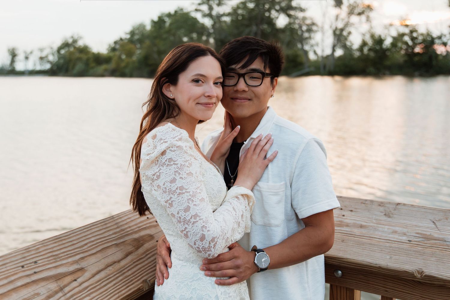 Smiling couple embracing on a wooden dock with a calm lake and sunset in the background.