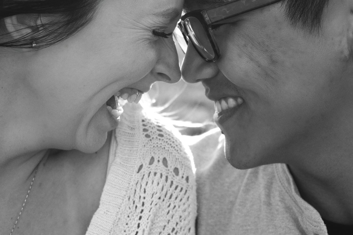 Black and white photo of a smiling couple sharing a romantic nose-to-nose moment, one wearing glasses.