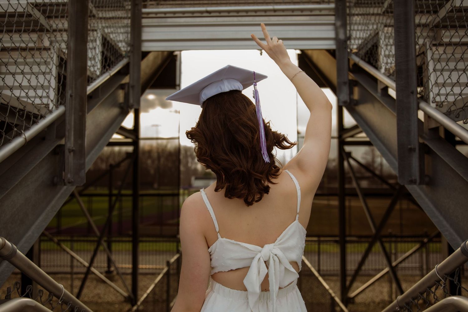 A graduate in a white dress and cap raises her hand in celebration on an outdoor metal staircase.