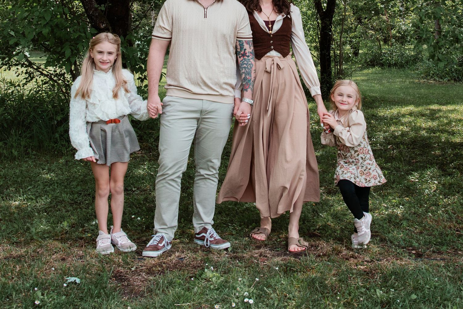 Family of four holding hands walking in a lush green park, parents with two young girls in casual outdoor attire.