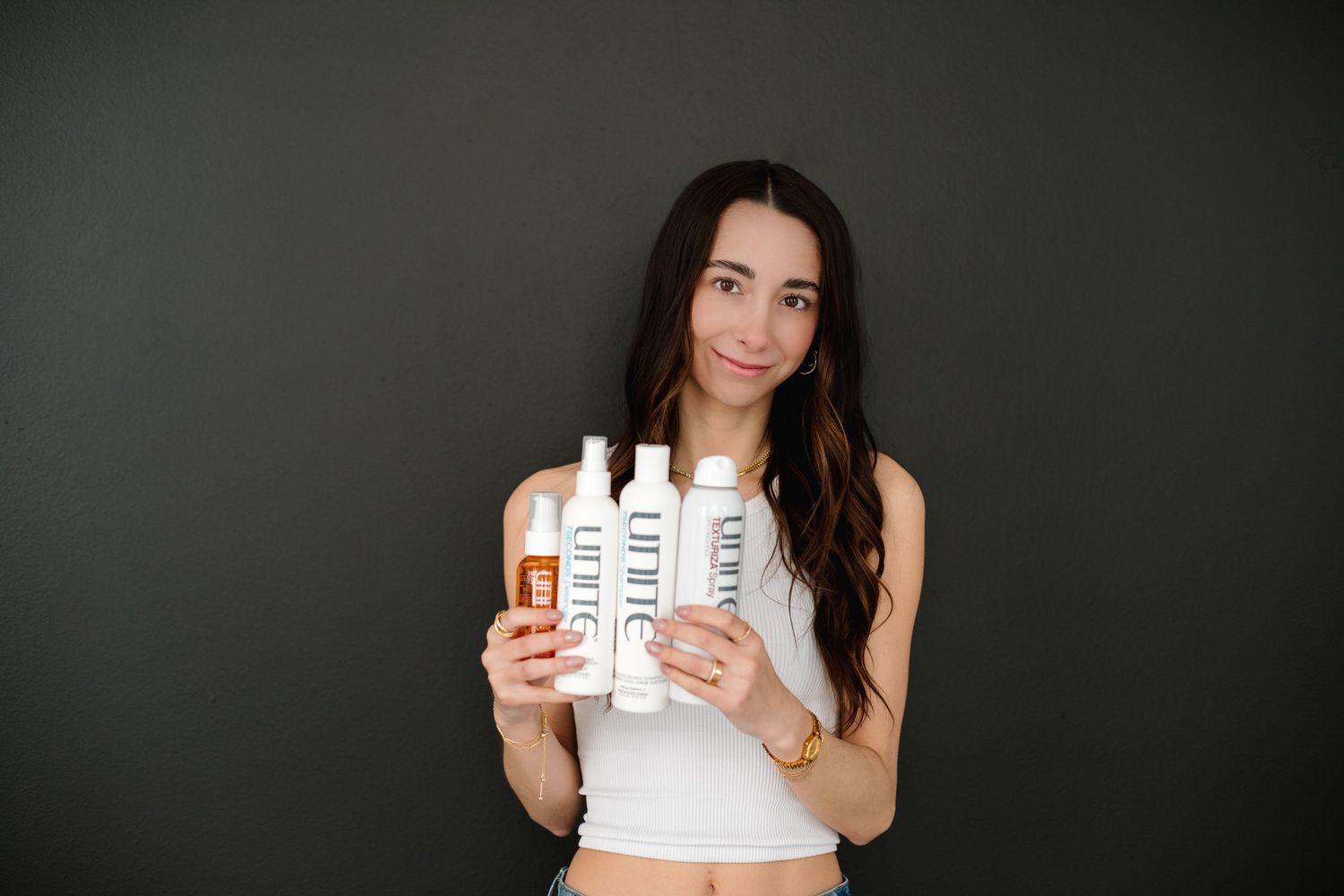 Brunette woman in white tank top holding Unite hair care product bottles against dark background.