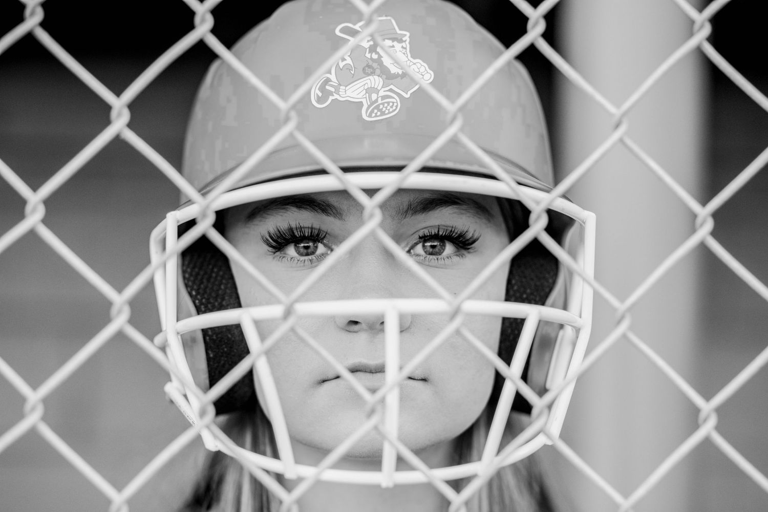 Black and white close-up of a female softball catcher staring through a chain-link fence in helmet.