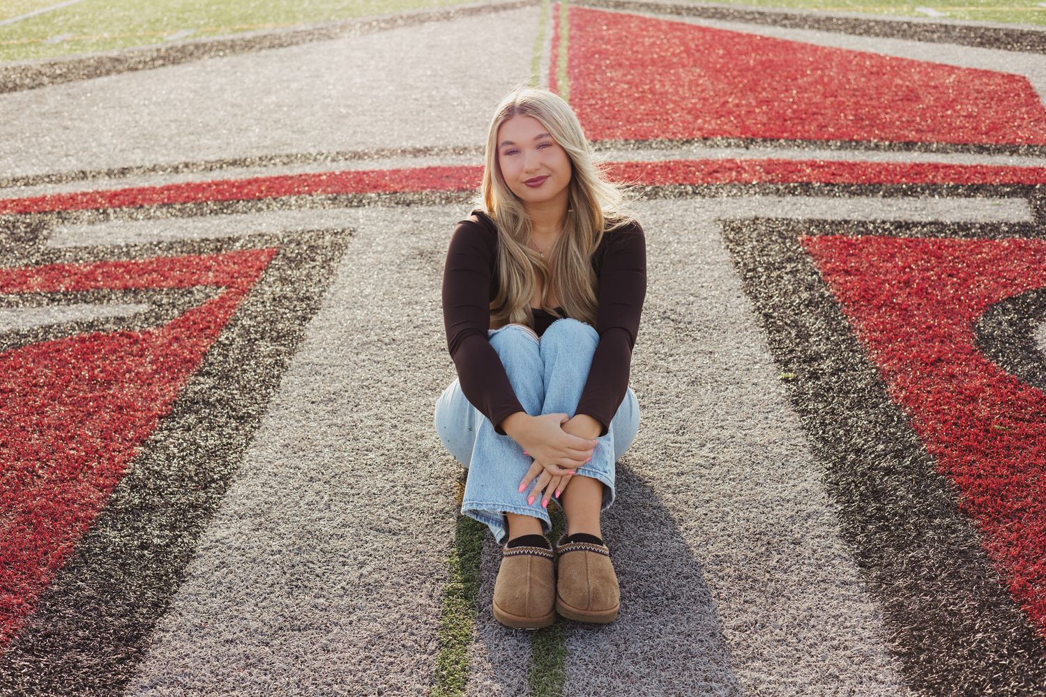 Blonde teenage girl in black sweater and jeans sitting on colorful school field turf.