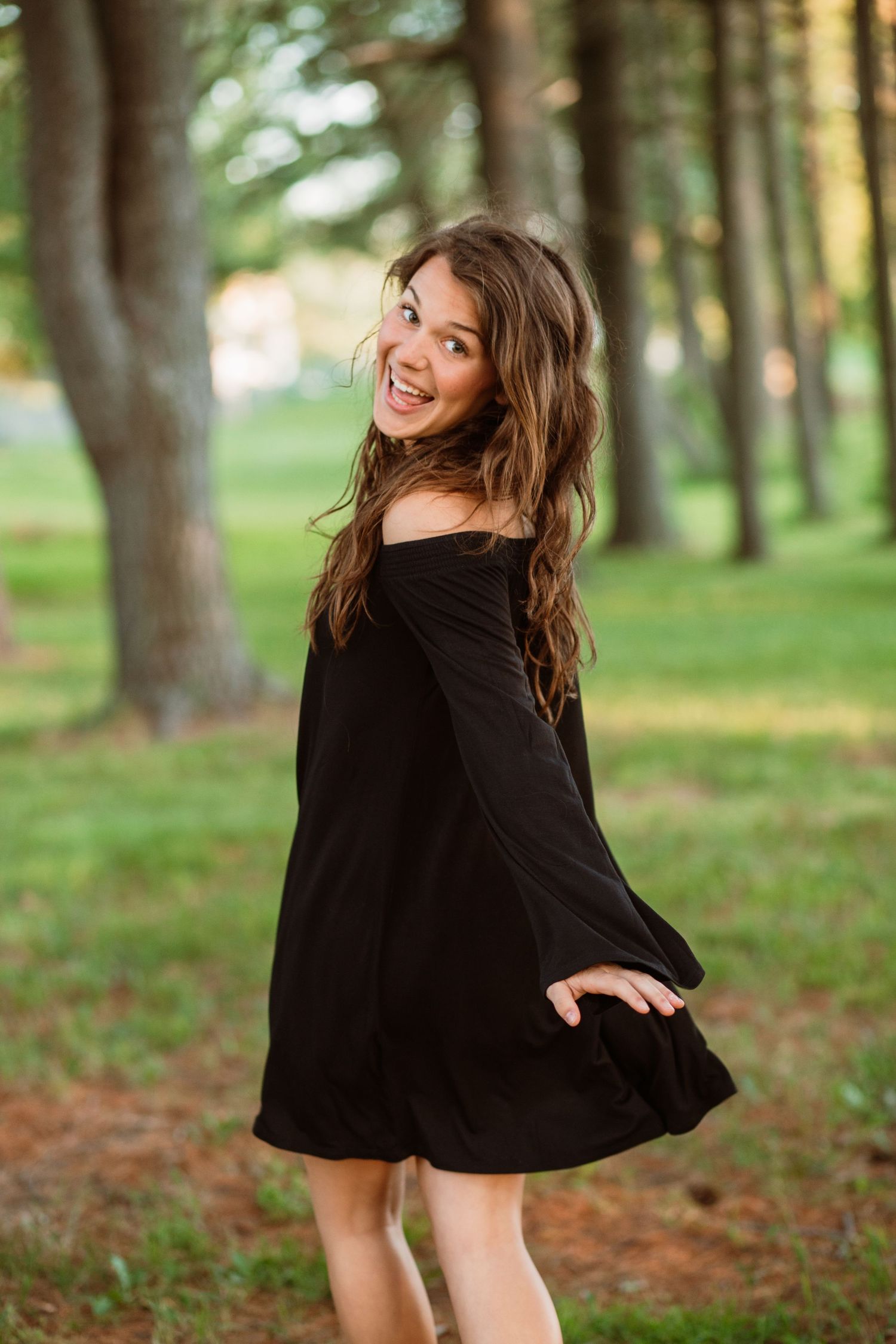 A young woman in a black off-shoulder dress smiles over her shoulder in a tree-lined park.