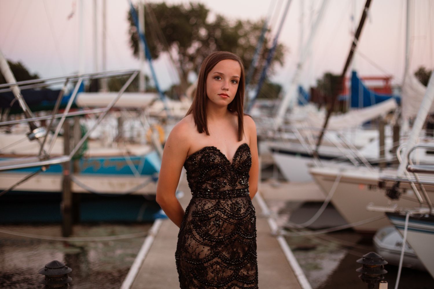 Teen girl in black strapless lace dress posing at marina with sailboats in background.