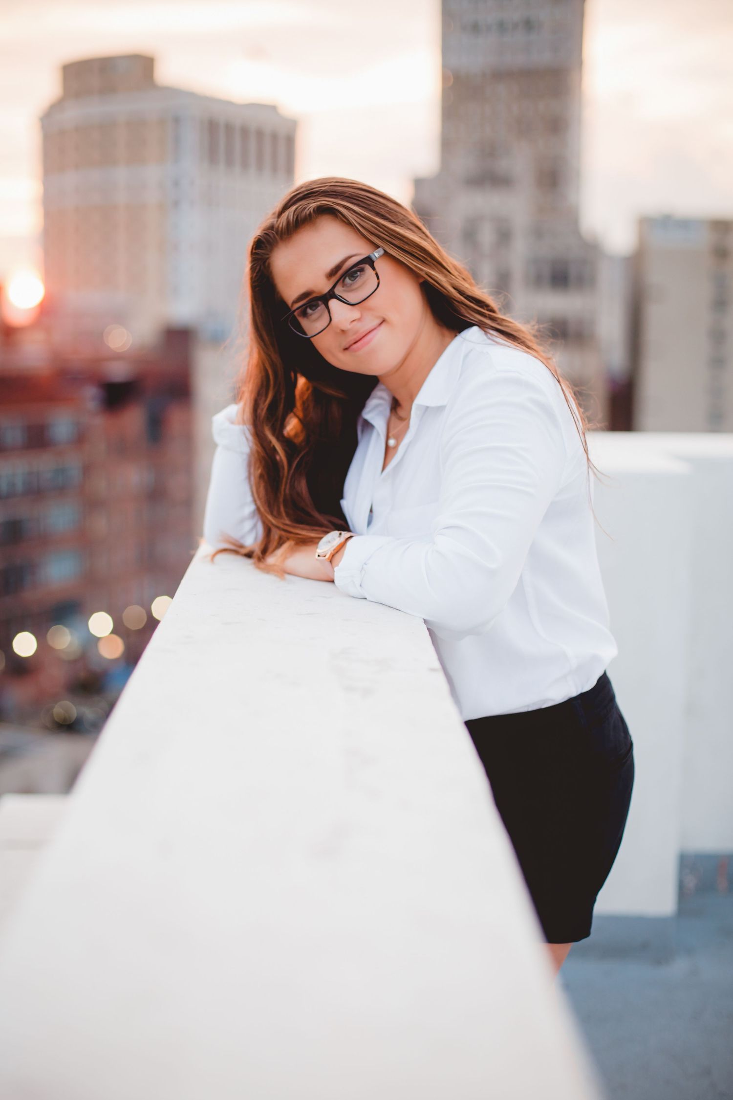 Young woman in white shirt and glasses leaning on rooftop railing at sunset.