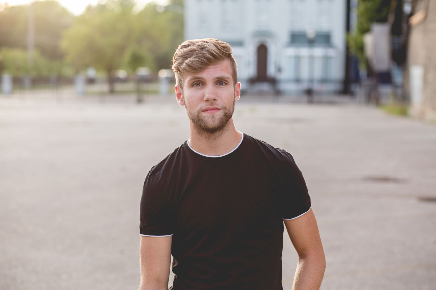 Young man with blonde beard wearing black t-shirt standing outdoors in urban setting during golden hour.