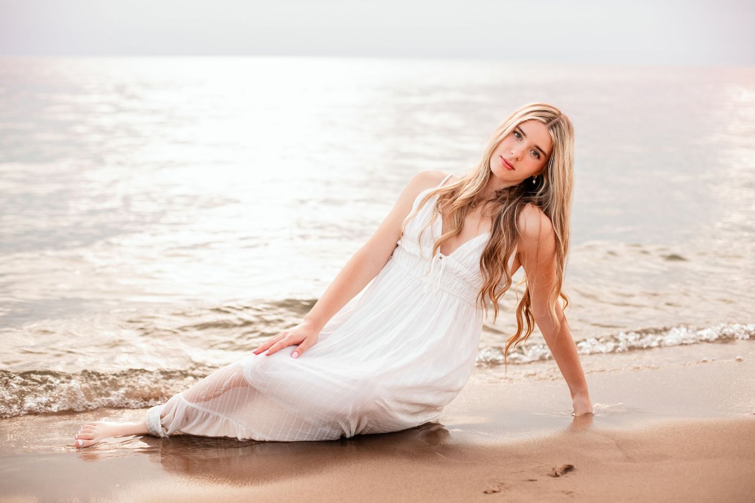 Young woman in white dress sitting on sandy beach shore with gentle ocean waves in warm golden light.